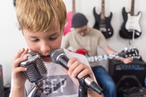 kid singing into 2 mics in front of a wall of guitars at Backbeat Music Academy