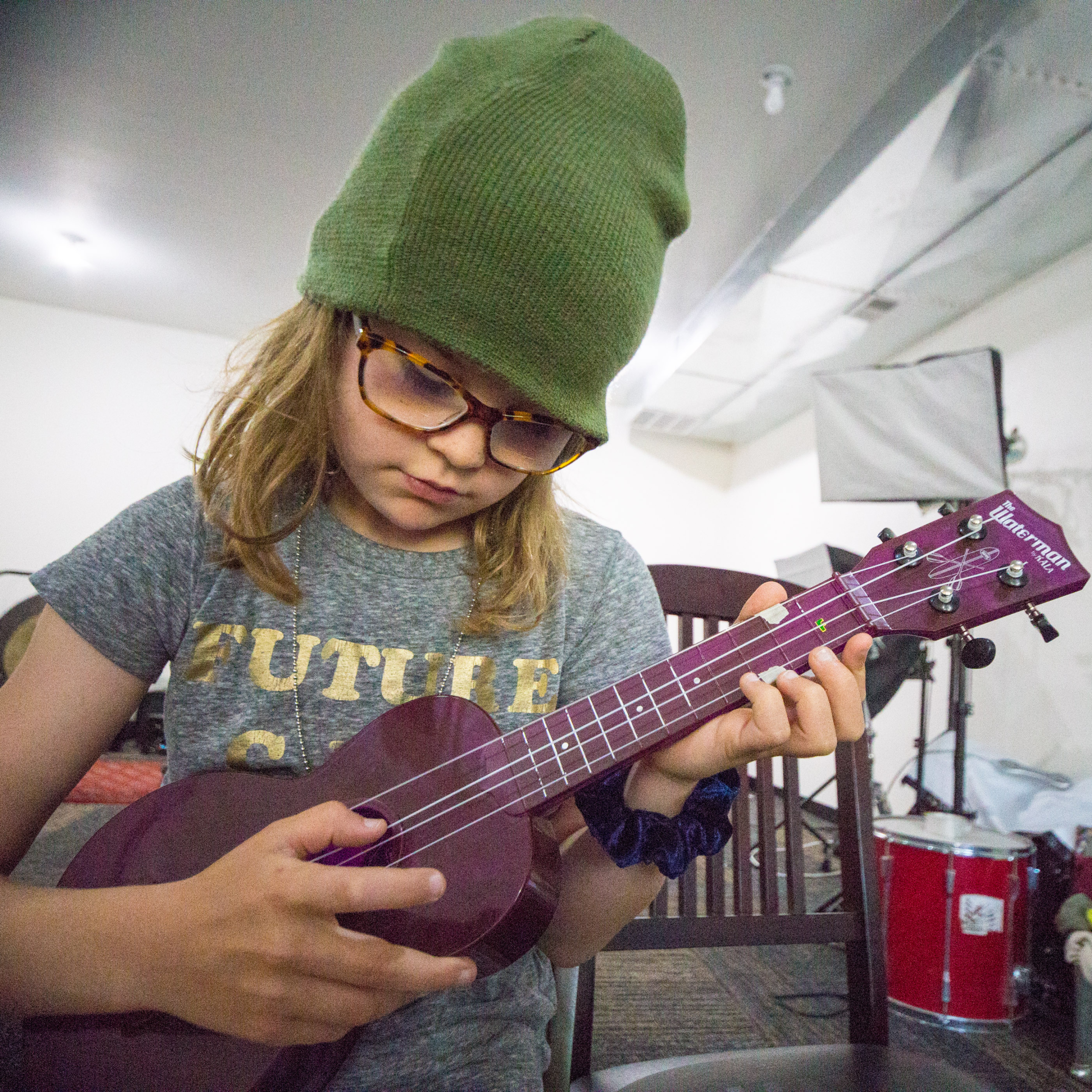 4 year kid strums purple ukulele in a white room. She is wearing a grey shirt and green beanie