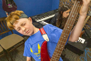 young bassist in a blue room with a piano behind him plays a red bass, bringing the headstock upwards and closing his eyes