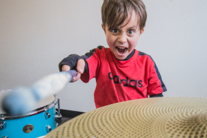 5 year old kid points a blue drumstick at the camera with an intense look on his face. He is sitting down on a blue kit wearing a red and black longsleeve adidas shirt