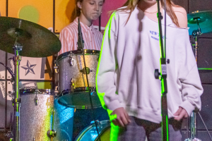 Young red headed 12 year old girl singing on a stage with a disappointed 14 year old drummer female playing a silver kit in the background