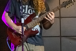 17-year-old male with long hair in front of his face playing a red guitar, standing behind a microphone with purple lighting coming down from his right